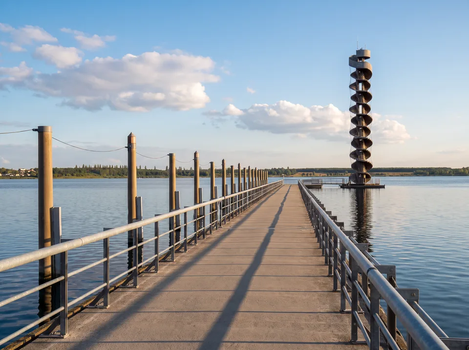 Seebrücke mit Geländer, die zum spiralförmigen Pegelturm im Goitzschesee führt, bei Sonnenschein