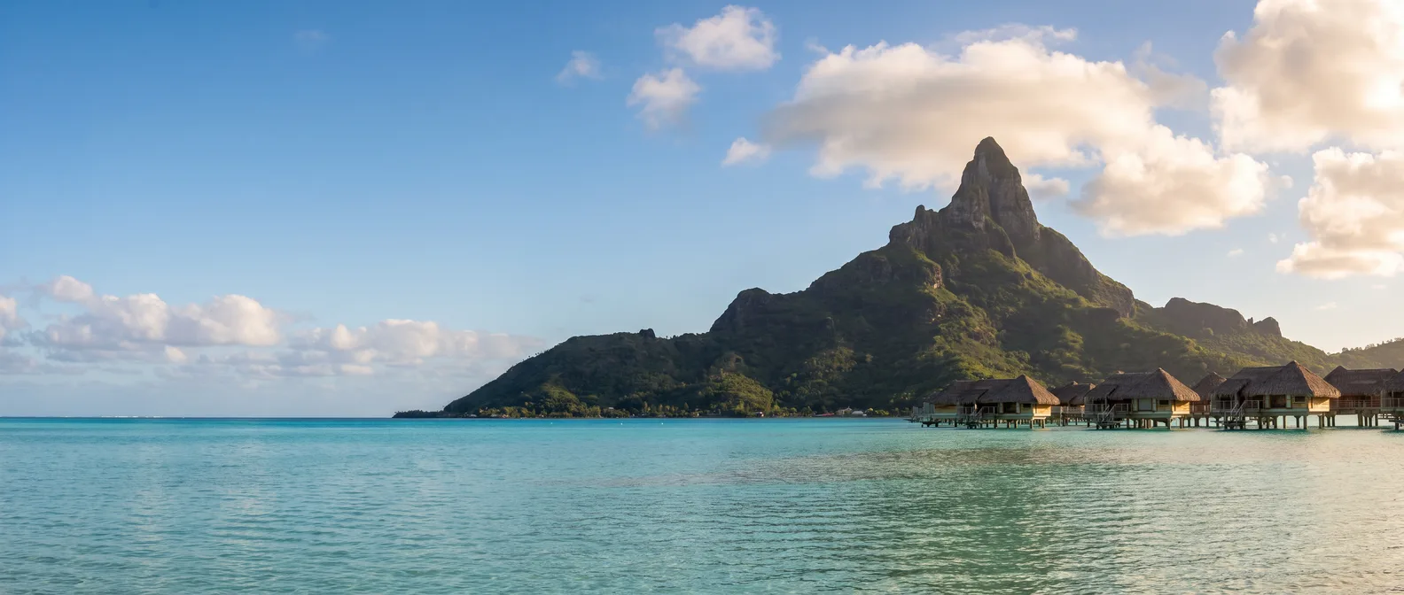 Der Mont Otemanu auf Bora Bora über der türkisfarbenen Lagune mit Überwasser-Bungalows auf Stelzen im Abendlicht