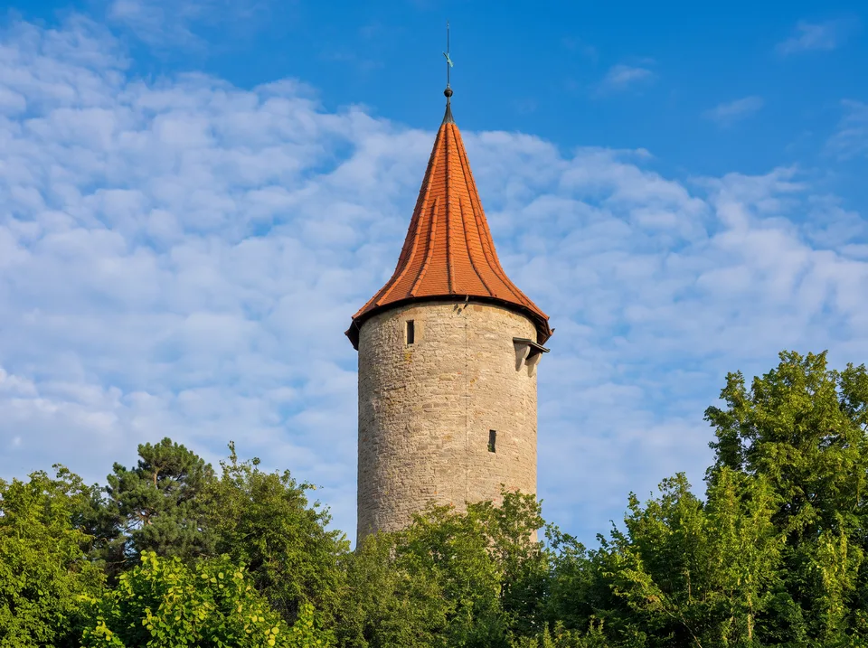 Der runde Diebsturm der Crailsheimer Stadtmauer aus Sandstein mit spitzem rotem Ziegeldach über grünen Bäumen