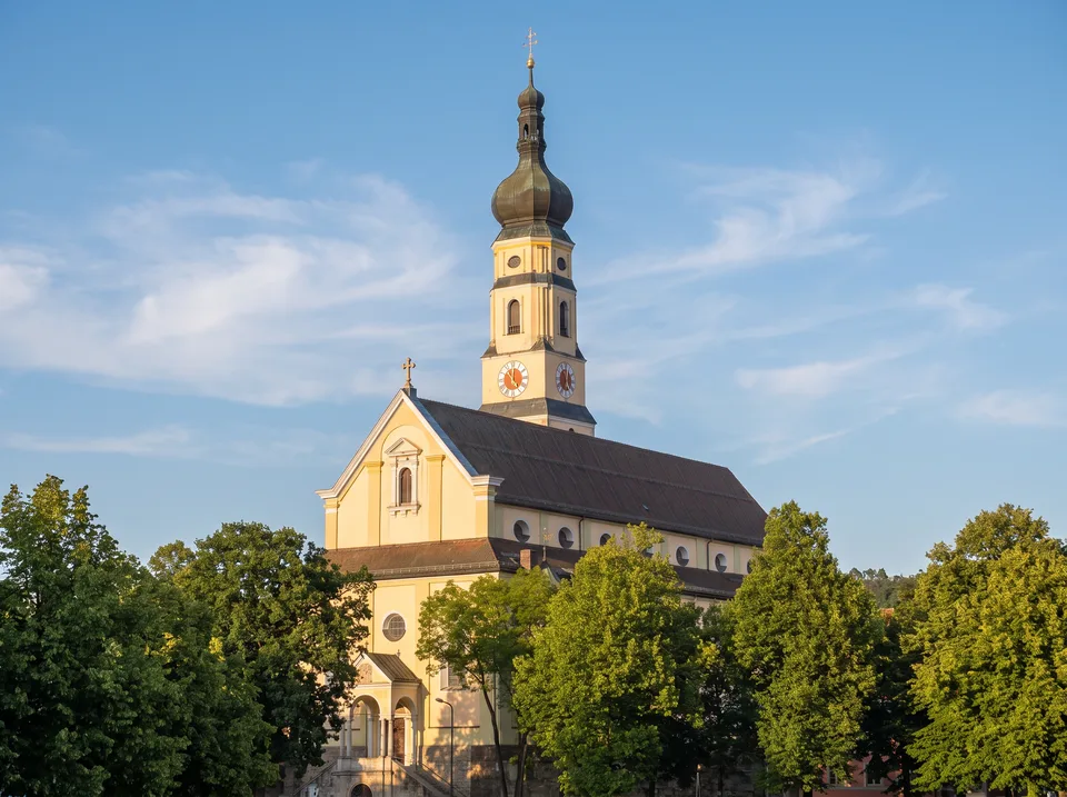 Die gelbe Stadtpfarrkirche Mariä Himmelfahrt in Deggendorf mit Uhrturm und Zwiebelhaube zwischen grünen Bäumen