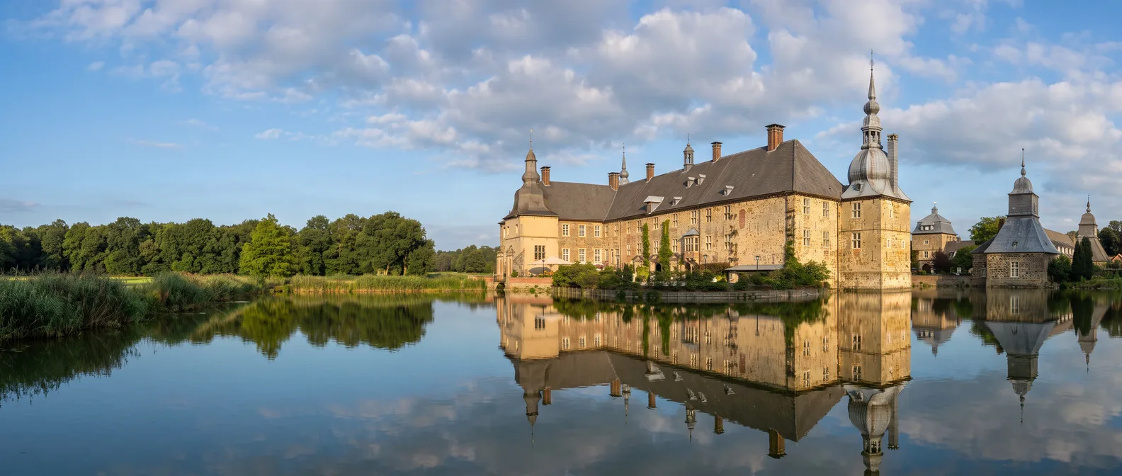 Das barocke Wasserschloss Lembeck mit Sandsteinfassade, Schieferdach und Turm mit geschwungener Haube spiegelt sich im Schlossteich unter blauem Himmel