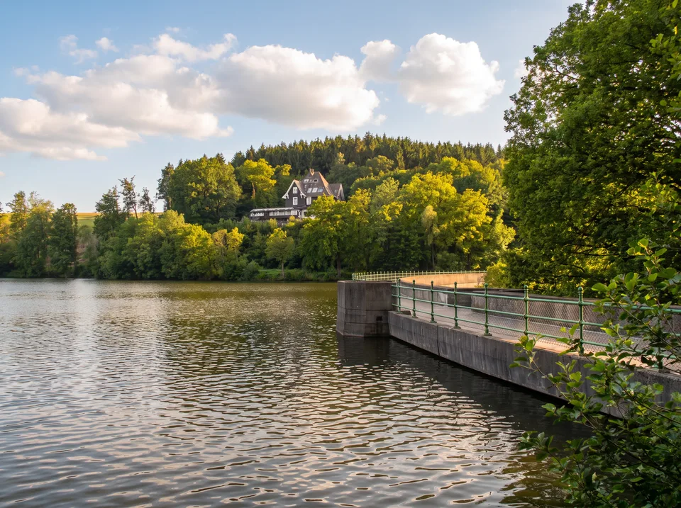 Die Heilenbecketalsperre mit Staumauer, grünem Eisengeländer und bewaldetem Hang im Sonnenlicht
