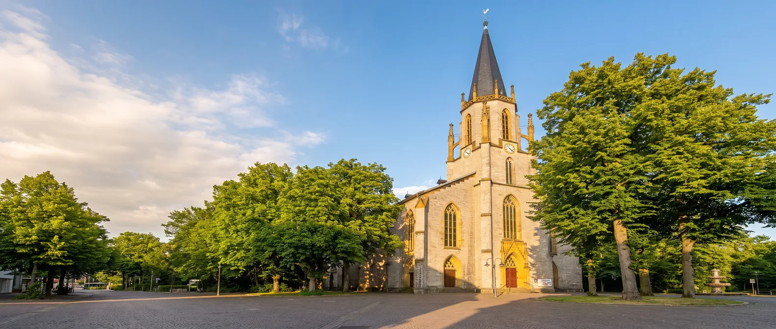 Neugotische Martin-Luther-Kirche in Gütersloh mit schlankem Westturm und spitzem Schieferhelm zwischen großen Bäumen im goldenen Spätsommerlicht