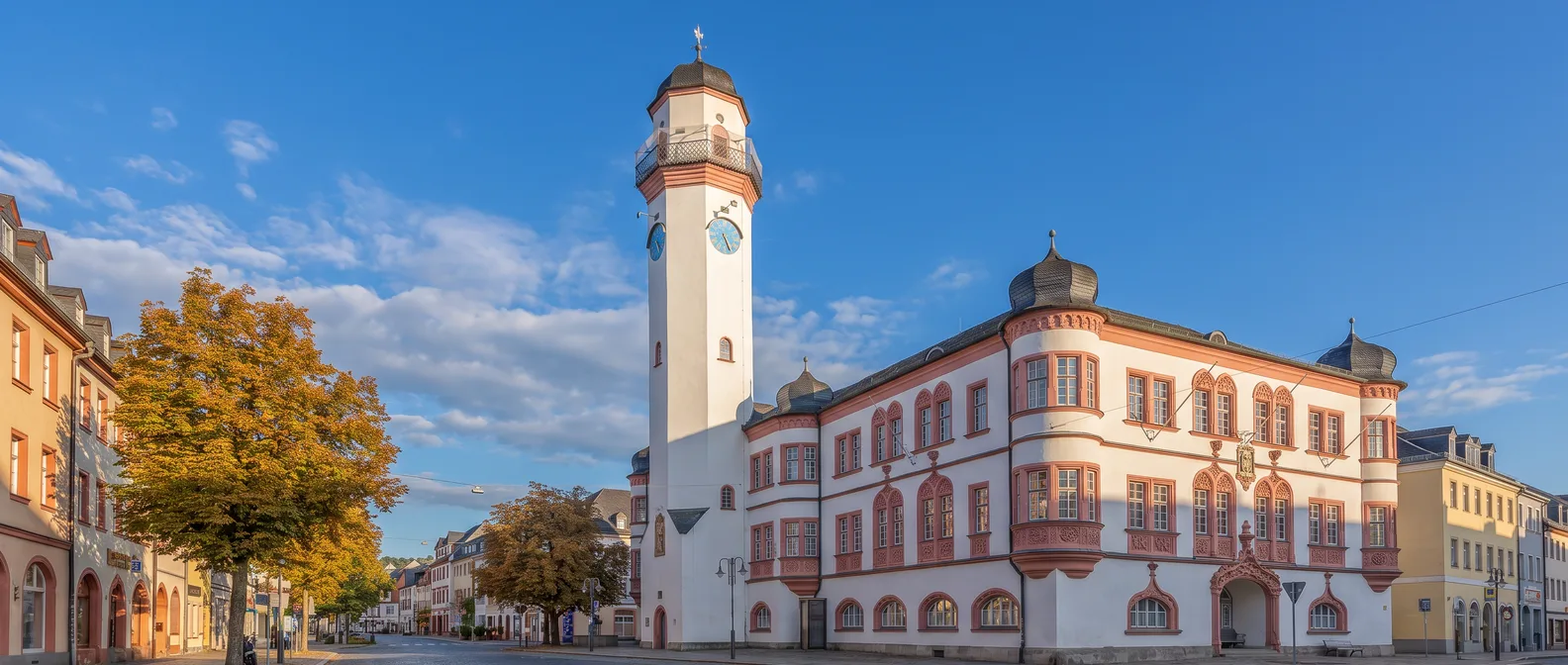 Das weiße Rathaus von Hof mit roten Fensterrahmen, Ecktürmchen und hohem achteckigem Uhrturm unter blauem Himmel