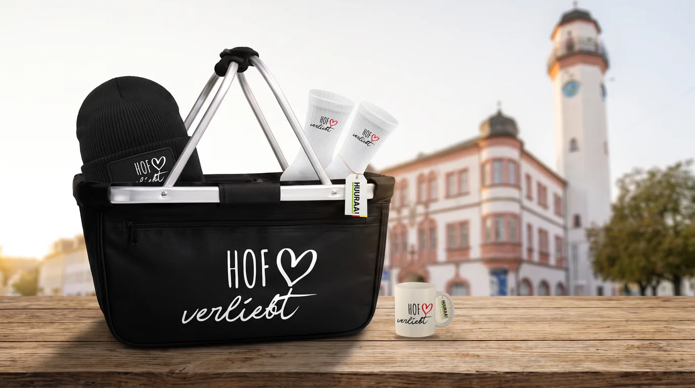Schwarzer Einkaufskorb Hof verliebt mit Beanie und Socken, daneben die Kaffeetasse, auf einem Holztisch vor dem Hofer Rathaus