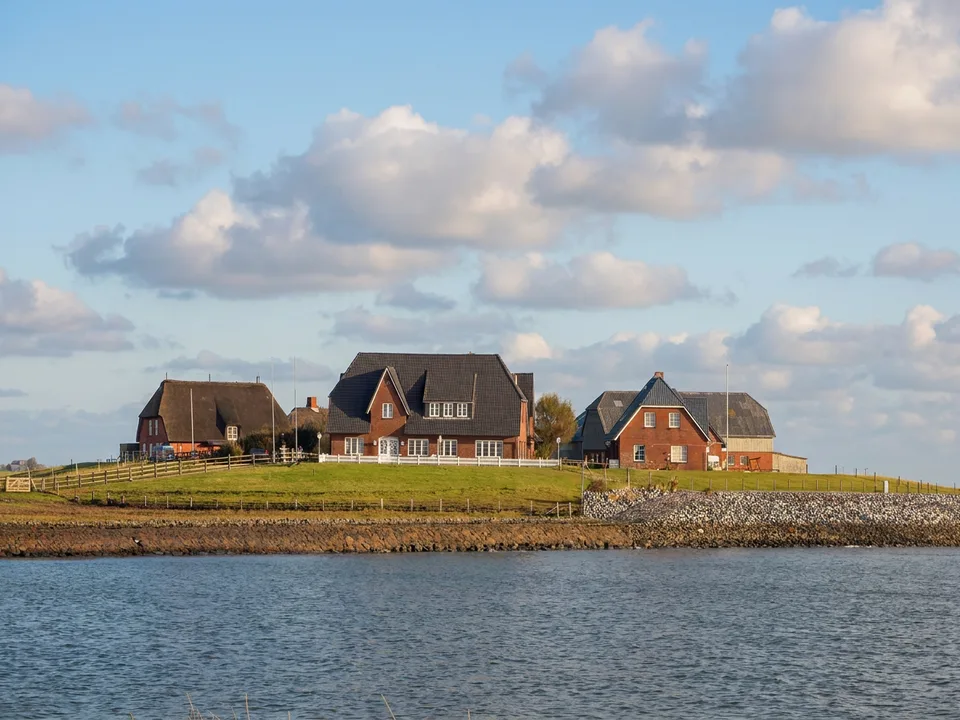 Backenswarft auf Hallig Hooge mit roten Backsteinhäusern auf der grünen Warft, davor Steinkante und Wasser