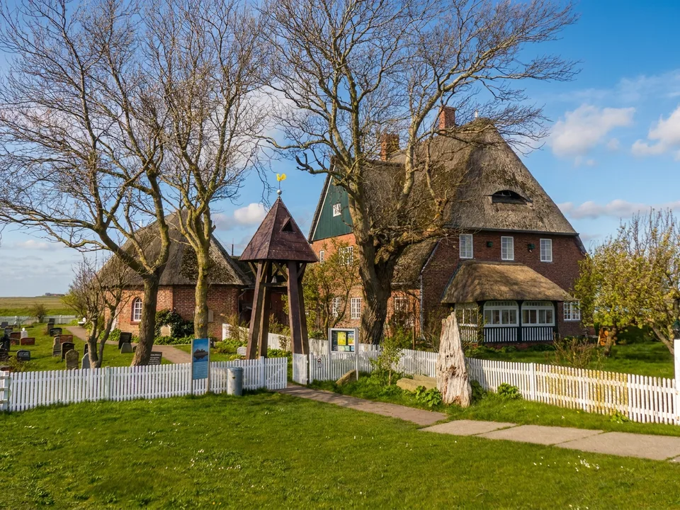 St.-Johannis-Kirche auf der Kirchwarft von Hooge mit hölzernem Glockenstapel, reetgedecktem Pfarrhaus und weißem Lattenzaun