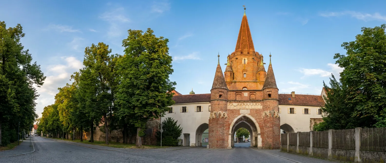 Das Kreuztor in Ingolstadt, ein gotisches Backsteintor mit spitzem Turmhelm und Ecktürmchen, im warmen Abendlicht