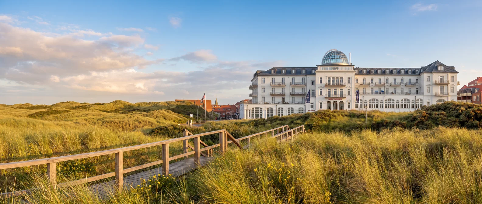 Weißes Strandhotel Kurhaus Juist mit Glaskuppel hinter Dünen mit Strandhafer und Holzsteg im Abendlicht