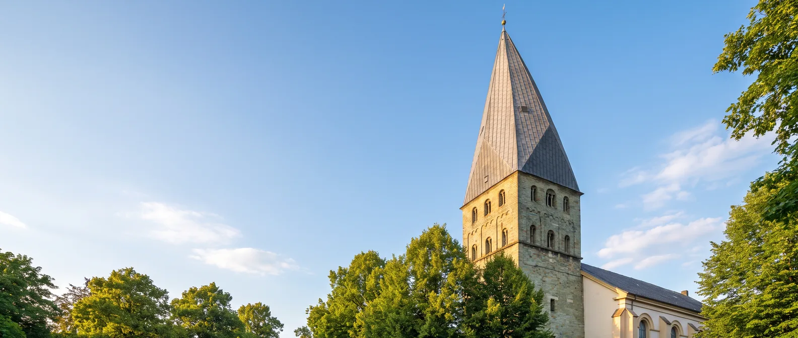 Der romanische Turm der Pauluskirche in Kamen mit seinem hohen, leicht verdrehten Schieferhelm über grünen Bäumen vor blauem Himmel