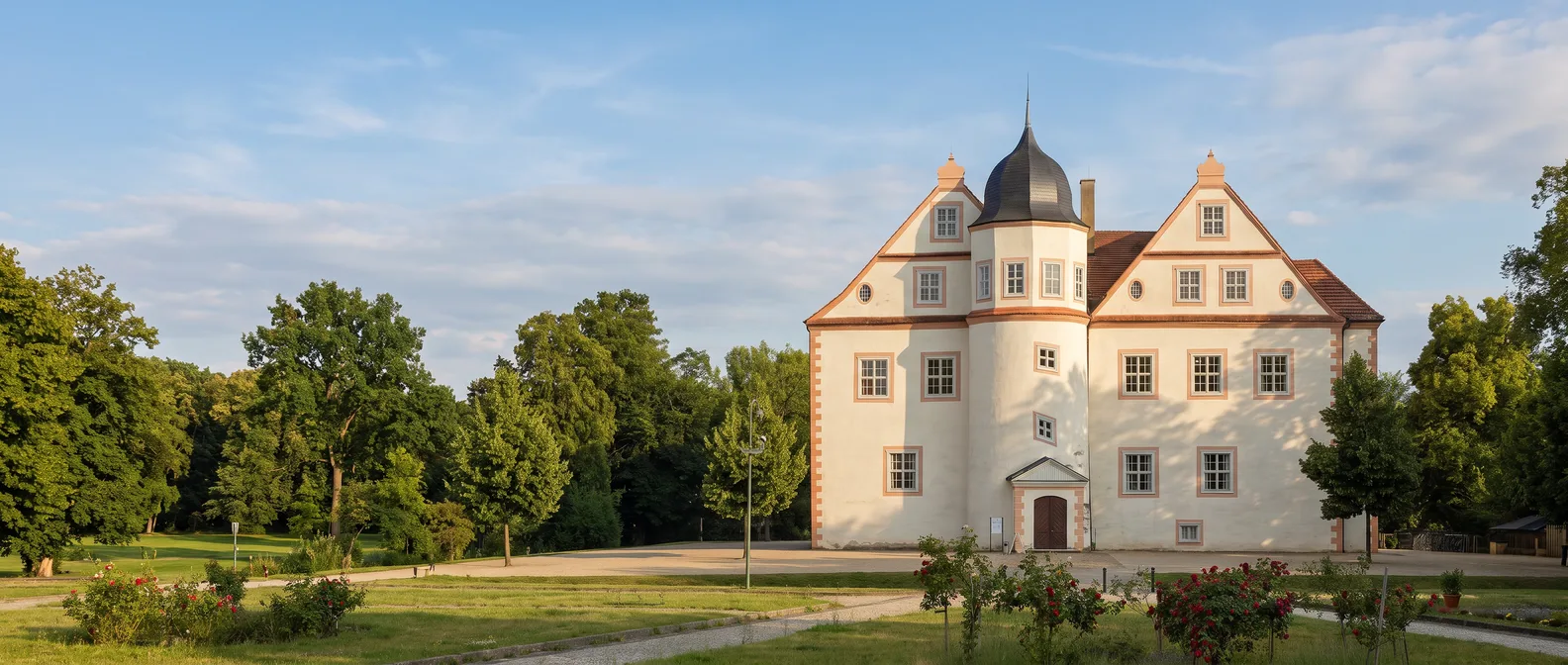 Das helle Schloss Königs Wusterhausen mit geschwungenen Giebeln und rundem Treppenturm mit dunkler Haube im warmen Abendlicht, links Bäume des Schlossparks