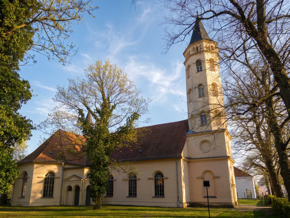 Die helle Kreuzkirche in Königs Wusterhausen mit rotem Ziegeldach, Rundbogenfenstern und schlankem Westturm zwischen hohen Bäumen