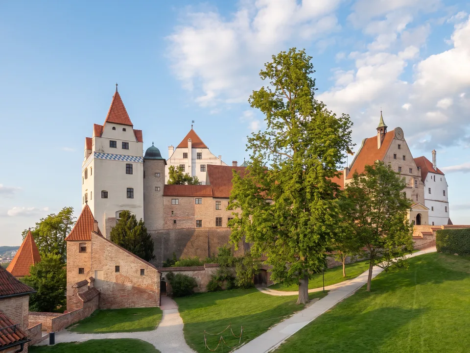 Burg Trausnitz mit weißem Turm, roten Ziegeldächern und Backsteinmauern über grünen Rasenflächen unter blauem Himmel