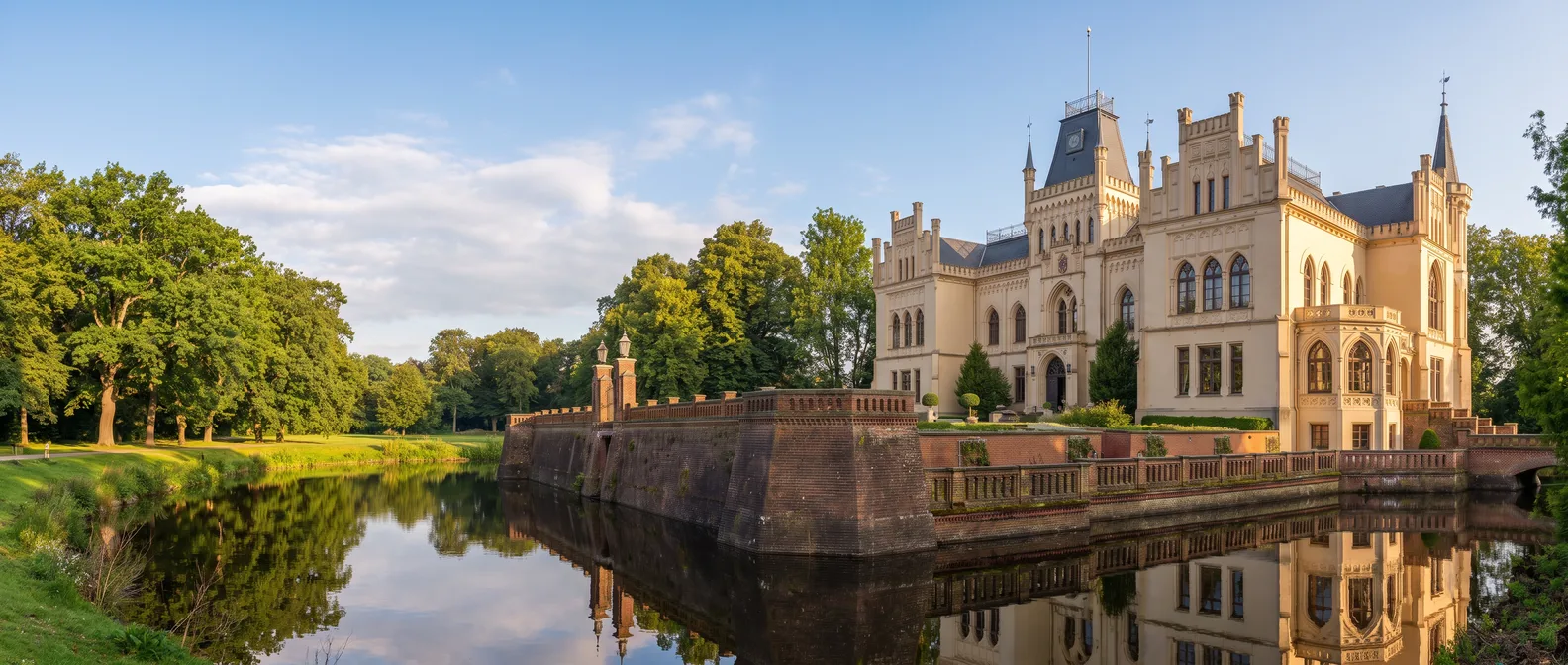 Die helle neugotische Evenburg in Leer-Loga mit Zinnengiebeln und Schieferturm hinter der Backsteinmauer, die sich im Schlossgraben spiegelt