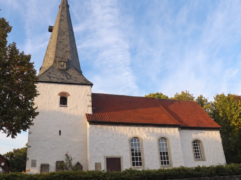 Kleine weiße Nikolauskirche in Lehrte mit schiefergedecktem Turmhelm und rotem Ziegeldach, davor eine Hecke unter blauem Himmel