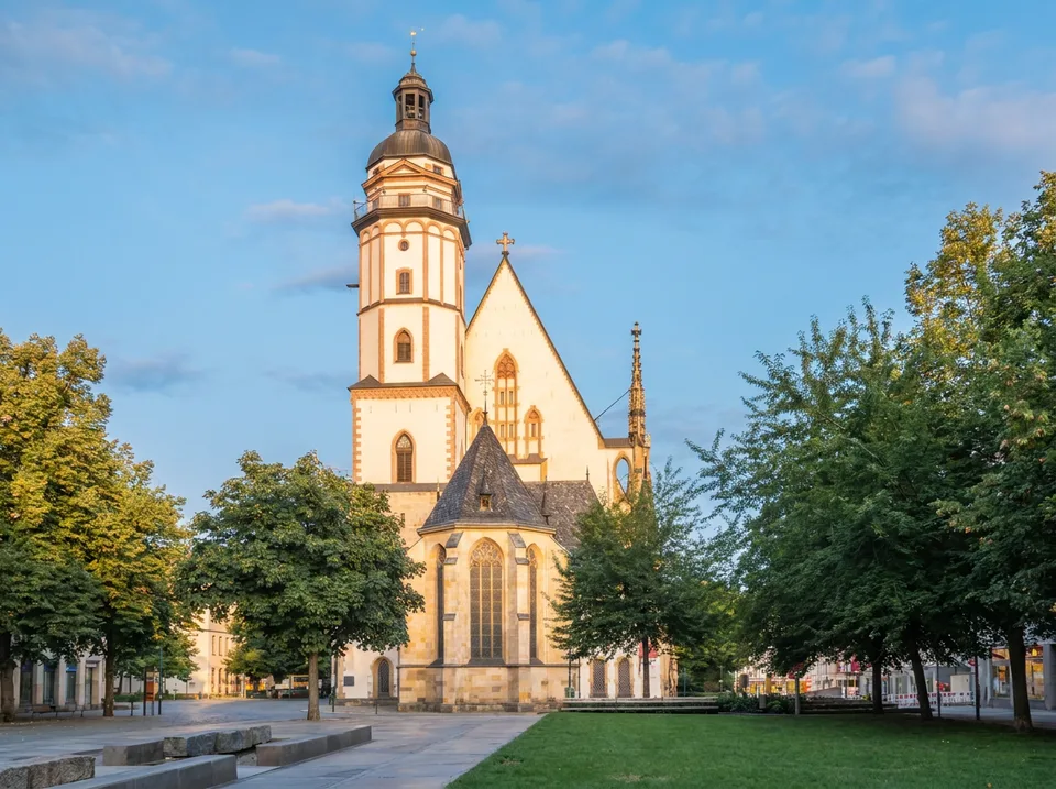 Die Thomaskirche in Leipzig mit hellem Turm, steilem Giebel und Chor zwischen grünen Bäumen
