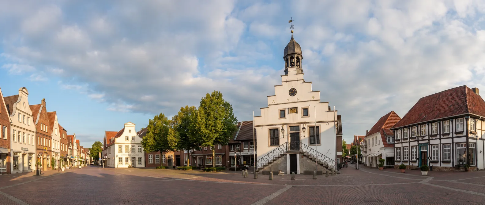 Das weiße historische Rathaus von Lingen (Ems) mit Treppengiebel, Uhr, Dachlaterne und doppelter Freitreppe am Marktplatz mit Giebel- und Fachwerkhäusern