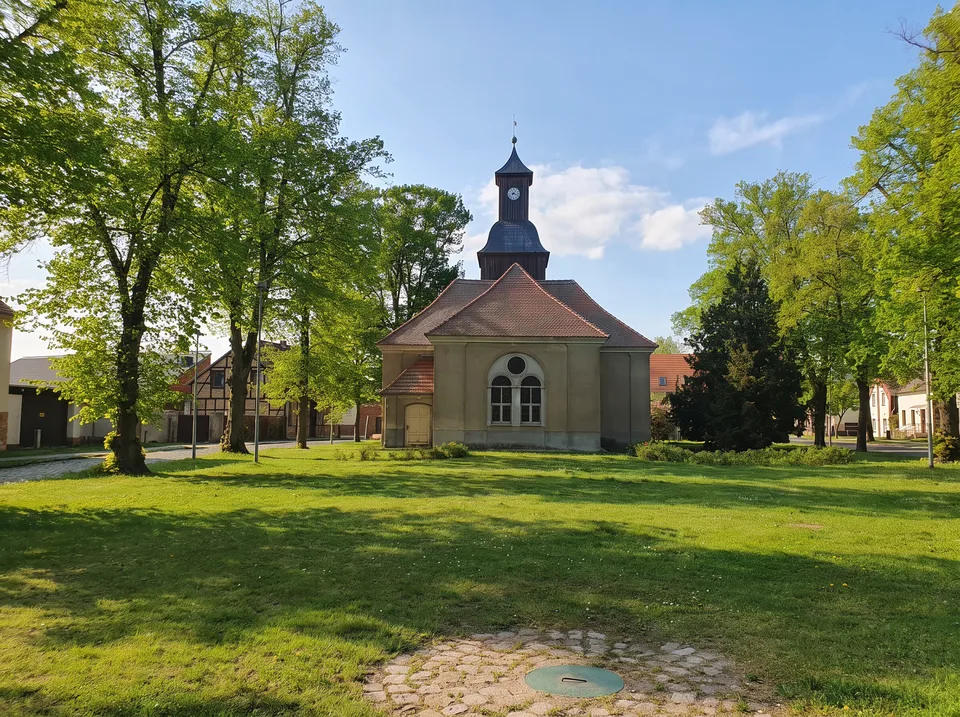 Die barocke Dorfkirche Groß Schulzendorf mit Turmlaterne und rotem Dach auf dem grünen Dorfanger unter hohen Bäumen