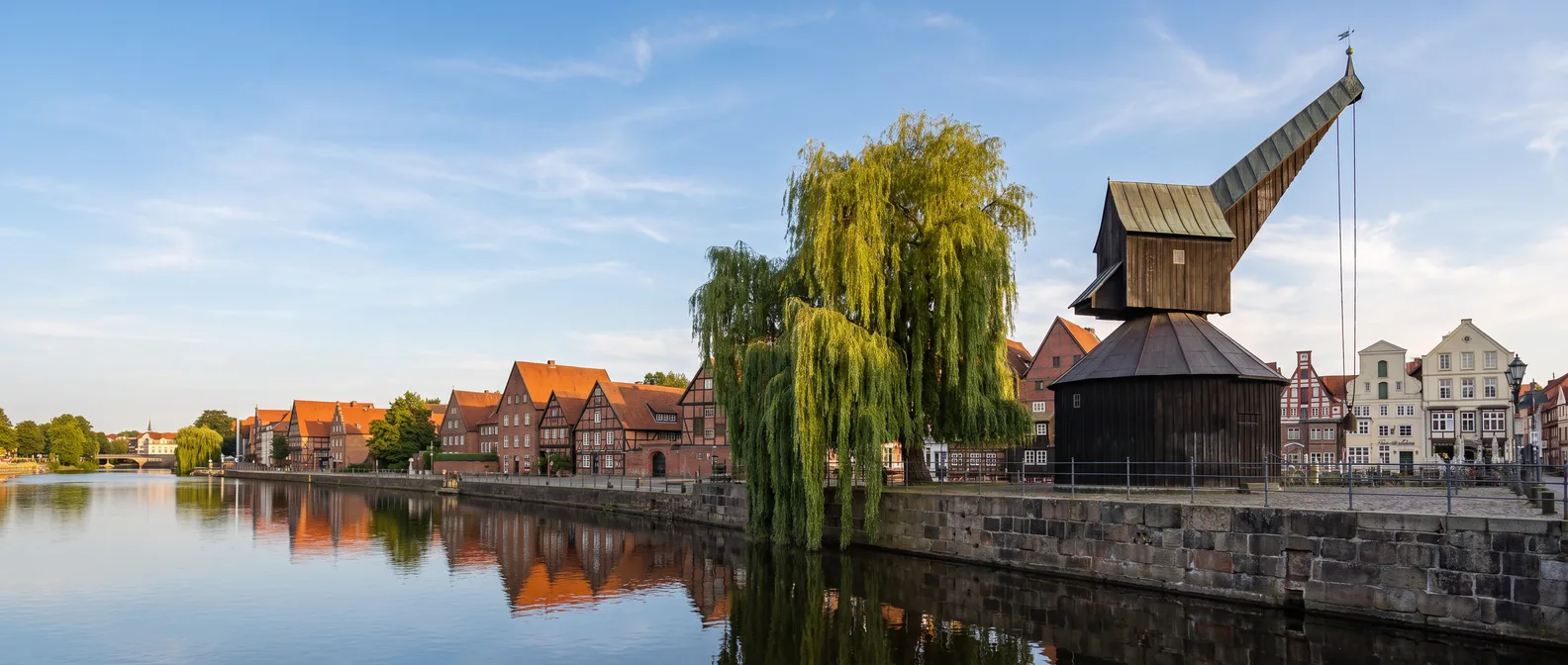 Der hölzerne Alte Kran mit Trauerweide an der Kaimauer des alten Ilmenauhafens in Lüneburg, links der ruhige Fluss mit historischen Giebelhäusern