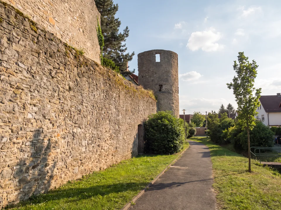 Die mittelalterliche Ringmauer in Maintal-Hochstadt aus Bruchstein mit rundem Turm, daneben ein Fußweg und junge Bäume