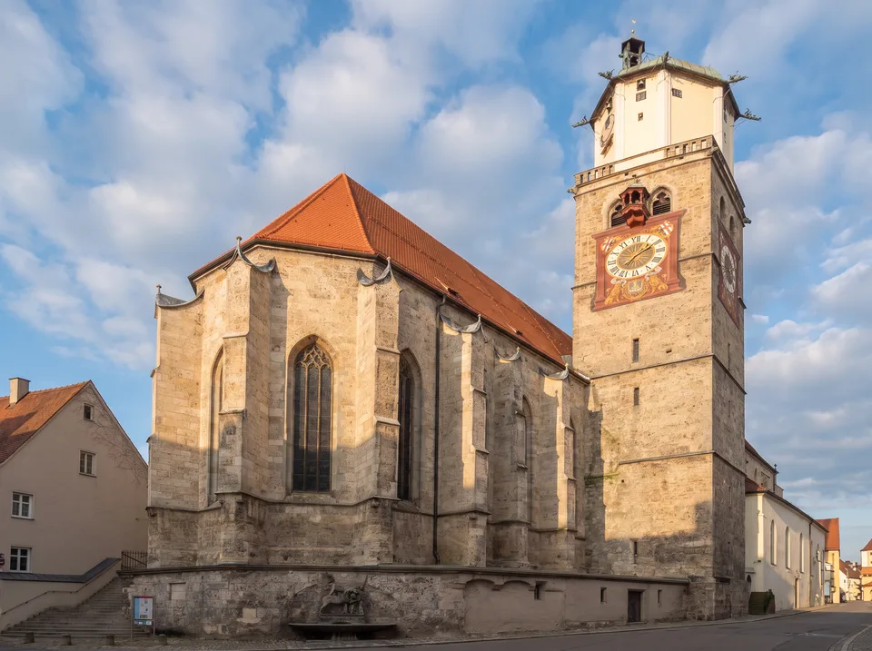 Gotischer Chor der Stadtpfarrkirche St. Martin in Memmingen aus hellem Sandstein mit rotem Ziegeldach, daneben der hohe Turm mit bemaltem Zifferblatt unter blauem Himmel