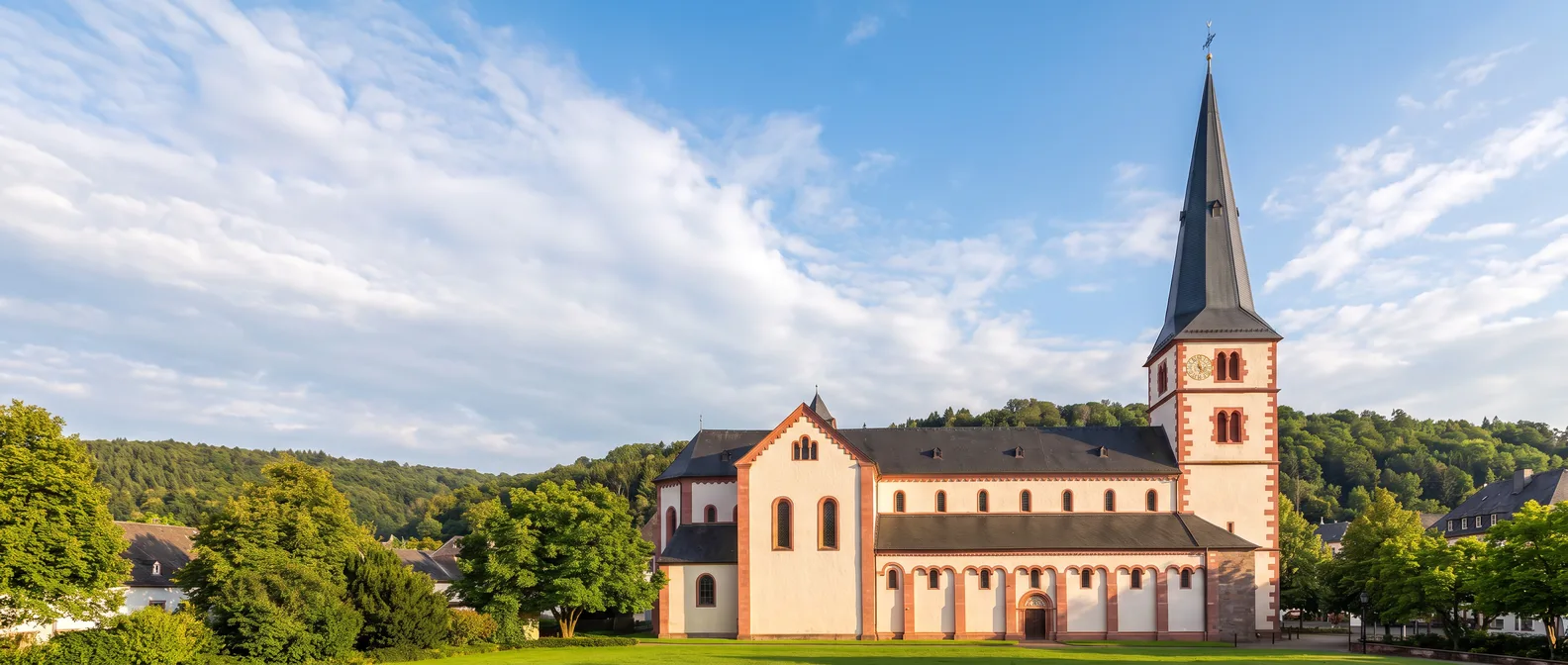 Die romanische Kirche St. Peter in Merzig mit weißen Mauern, roten Sandsteinkanten und spitzem Schieferturm vor bewaldeten Hügeln