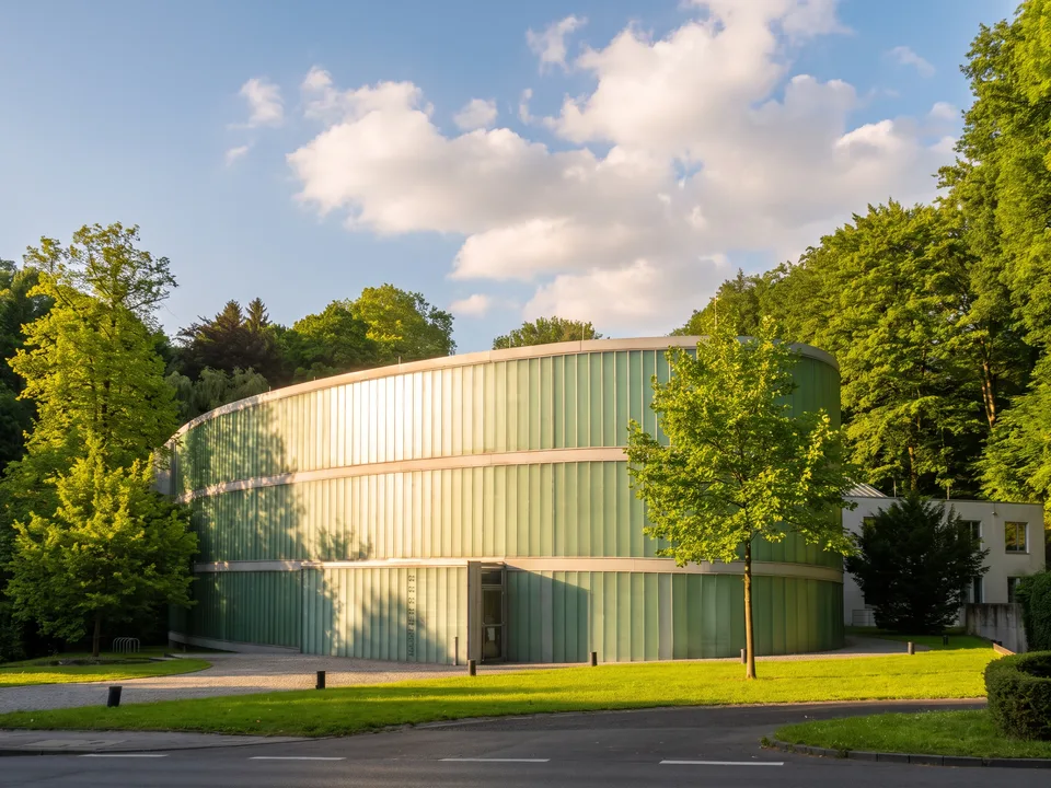 Das ovale Neanderthal Museum mit grünlich schimmernder Glasfassade zwischen Bäumen im Neandertal bei Mettmann