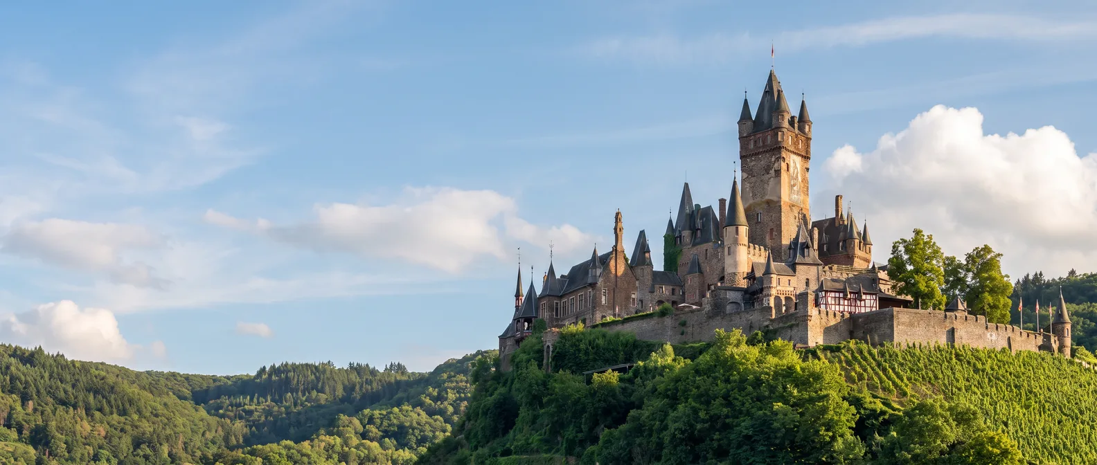 Reichsburg Cochem mit hohem Bergfried und spitzen Türmchen auf dem bewaldeten Bergkegel über den Weinbergen an der Mosel im Abendlicht