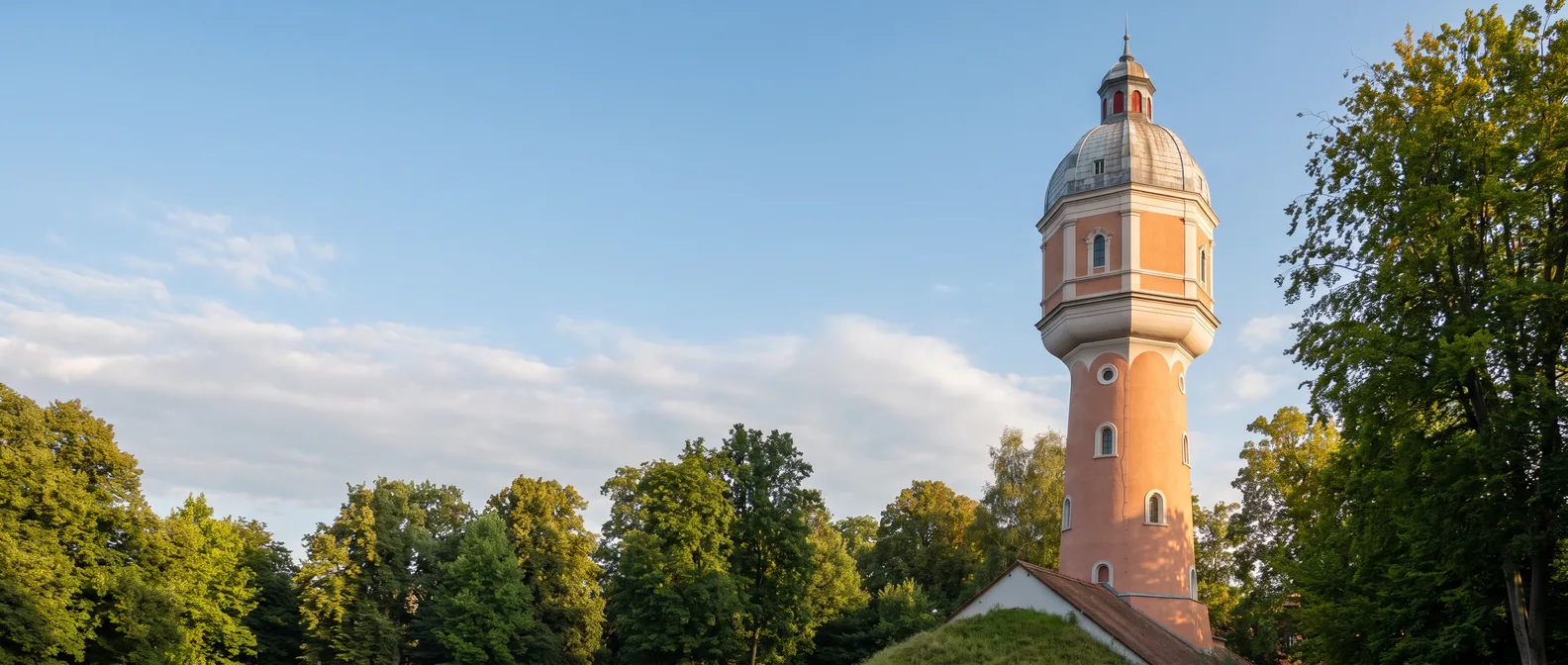 Der lachsrosa Wasserturm von Neu-Ulm mit achteckigem Behältergeschoss und grauer Haube über den grünen Bäumen des Kollmannsparks im Abendlicht