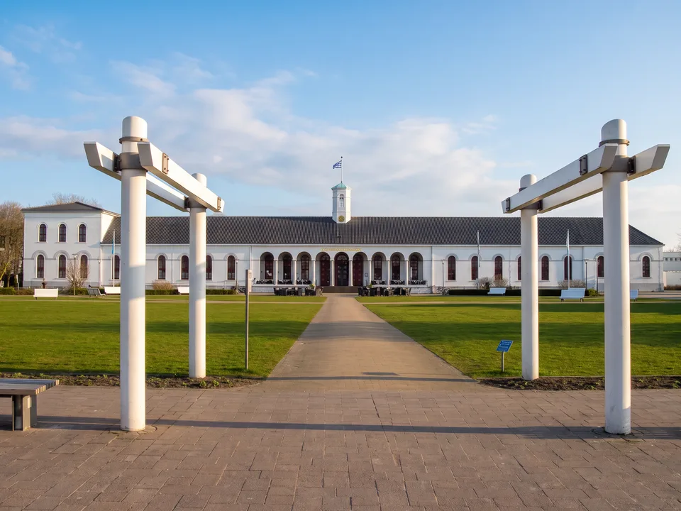 Weißes Conversationshaus Norderney mit Rundbogenarkaden und Dachreiter, davor Rasen und weiße Pergola-Pfeiler