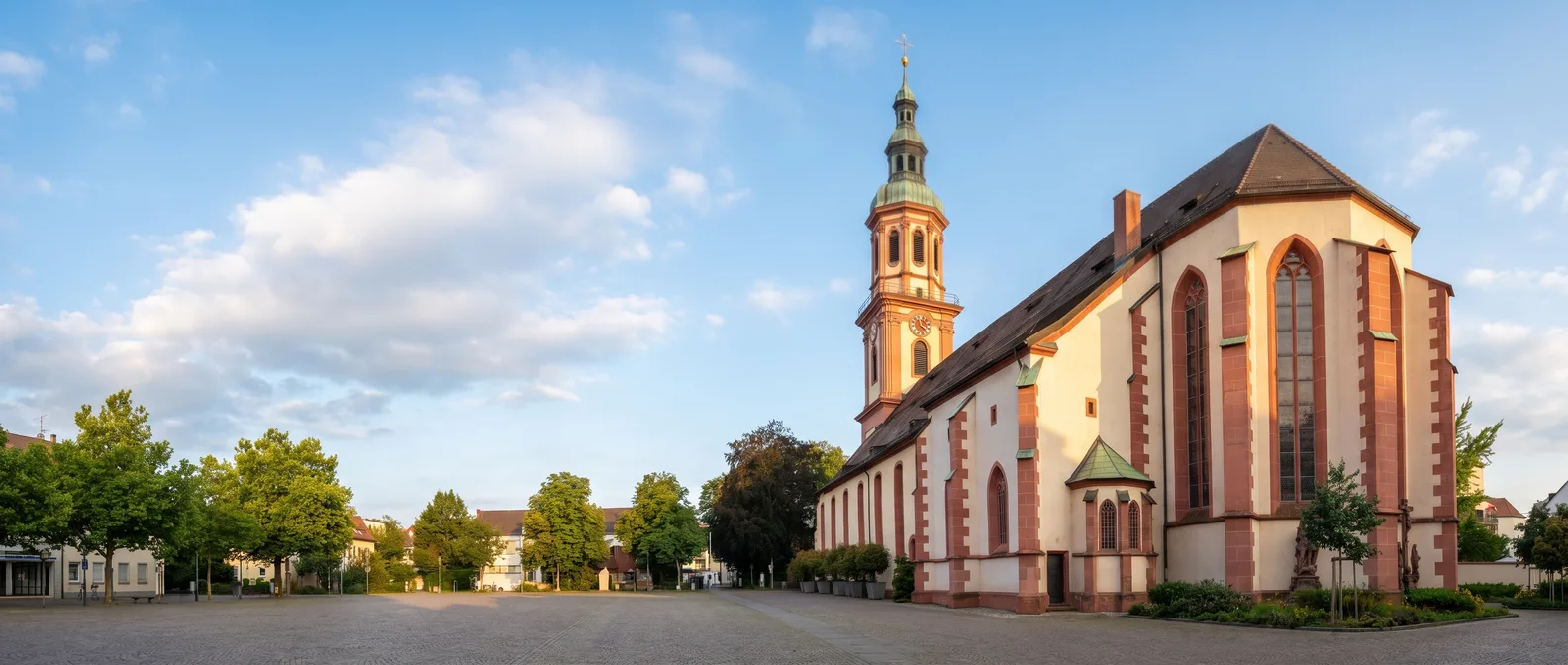 Die Heilig-Kreuz-Kirche in Offenburg mit hellen Wänden, roten Sandsteinpfeilern, gotischen Chorfenstern und barockem Turm mit grüner Haube am Abend