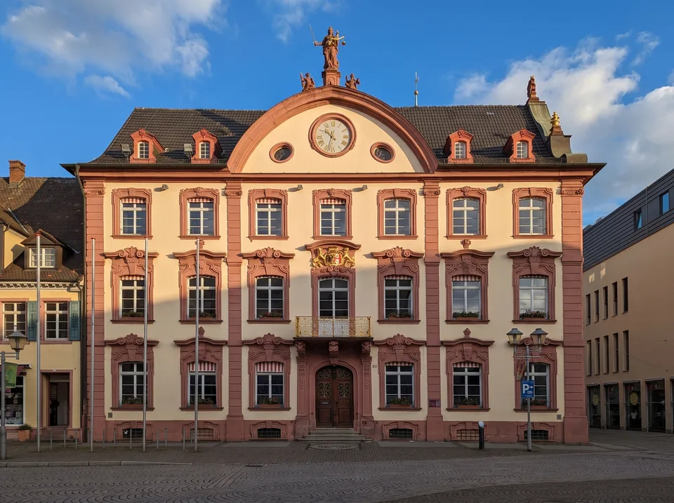 Das barocke historische Rathaus in Offenburg mit rosa-cremefarbener Fassade, rotem Sandstein, geschwungenem Mittelgiebel mit Uhr und Justitia-Figur