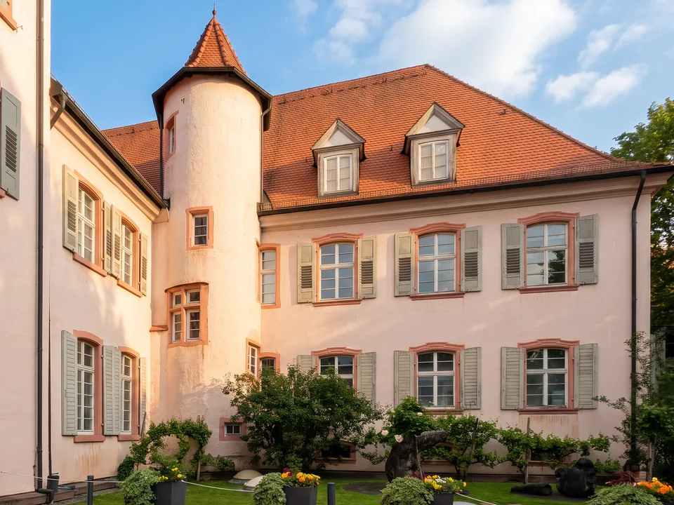 Das rosafarbene Ritterhaus in Offenburg mit rundem Treppenturm, roten Ziegeldächern, Gauben und grauen Fensterläden über einem Garten