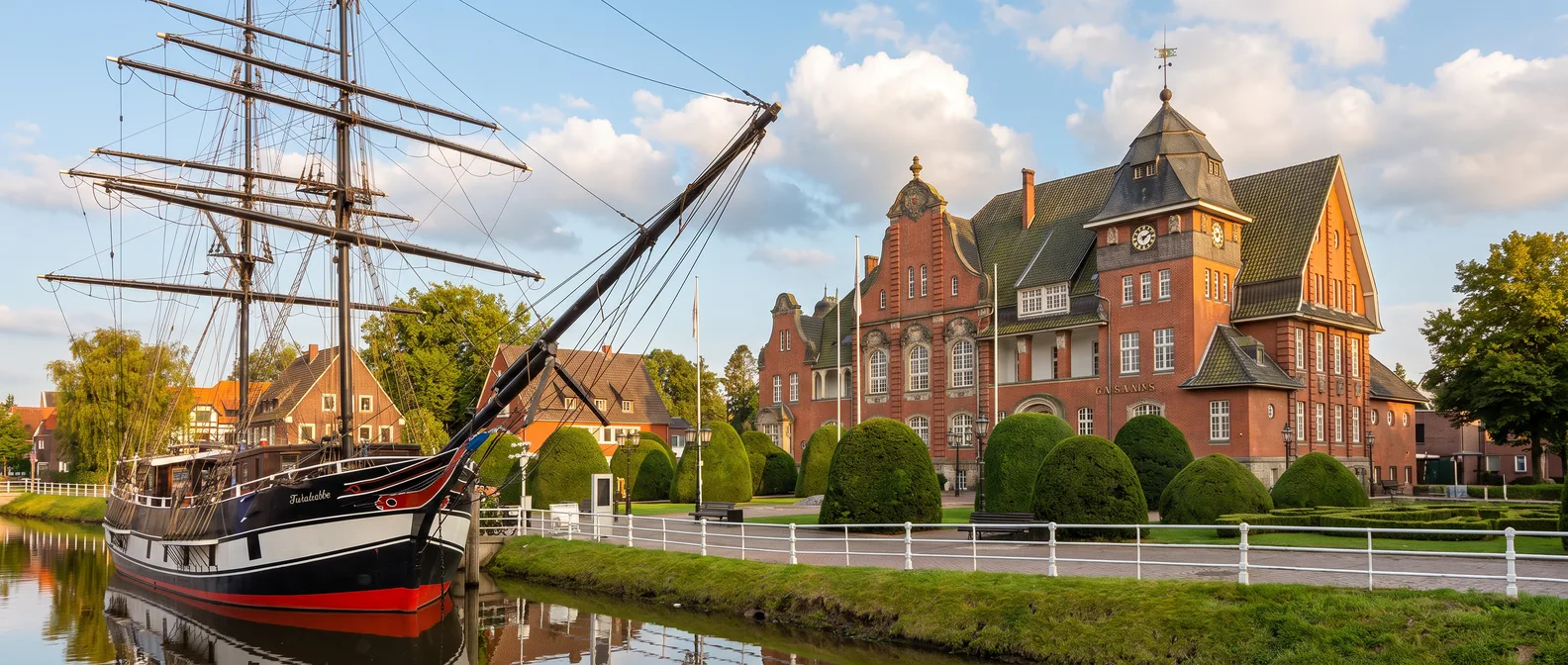Das neobarocke Rathaus von Papenburg mit Uhrturm hinter gestutzten Buchsbäumen, links im Kanal der Nachbau der Brigg Friederike