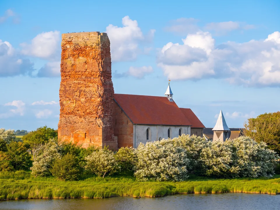 Turmruine der Alten Kirche St. Salvator auf Pellworm aus rotem Backstein neben dem weißen Kirchenschiff mit rotem Dach, davor blühende Büsche und Wasser