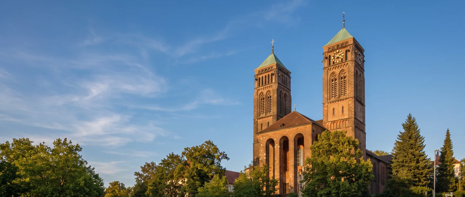Die Kirche St. Pirmin in Pirmasens aus rötlichem Sandstein mit zwei Türmen und grünen Kupferdächern im warmen Abendlicht, links Bäume und blauer Himmel