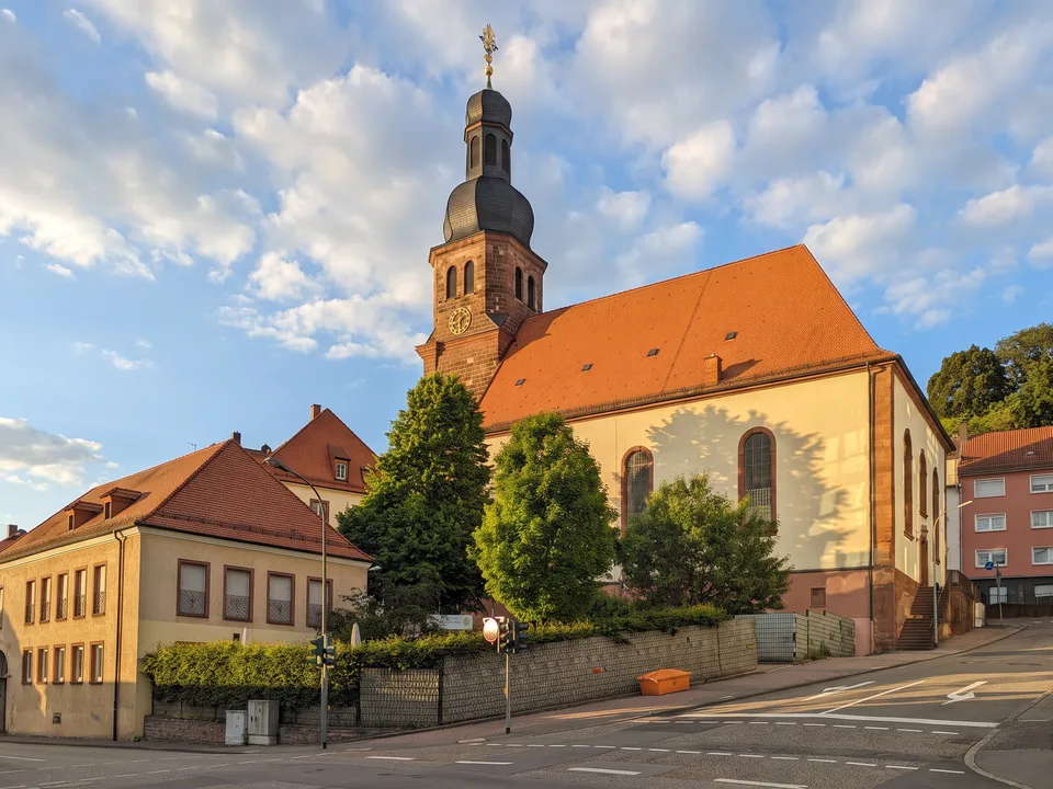 Die weiße Lutherkirche in Pirmasens mit orangerotem Ziegeldach und Sandsteinturm mit dunkler Haube, davor Bäume und Häuser