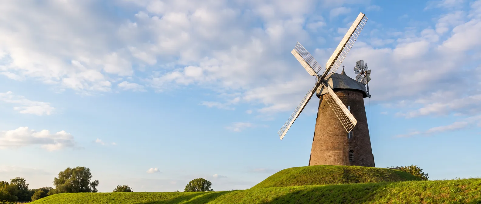 Die Stommelner Windmühle mit weißen Flügeln auf ihrem grünen Hügel in Pulheim-Stommeln unter blauem Himmel