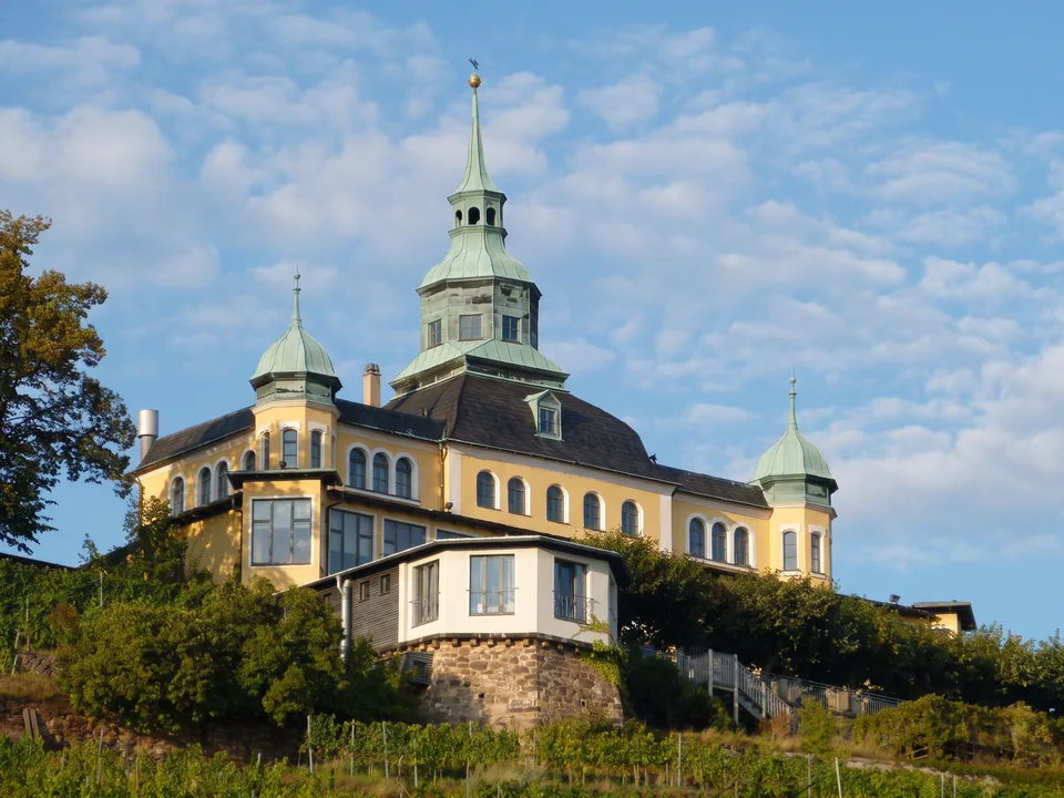 Das gelbe Spitzhaus in Radebeul mit grünem Kupferturm und Ecktürmchen auf der Hangkante über den Weinbergen