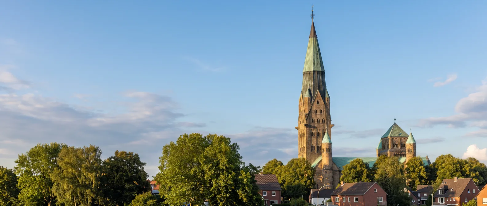 Die neuromanische Basilika St. Antonius in Rheine mit ihrem hohen Turm und grünem Kupferhelm über Bäumen und Backsteinhäusern im Abendlicht