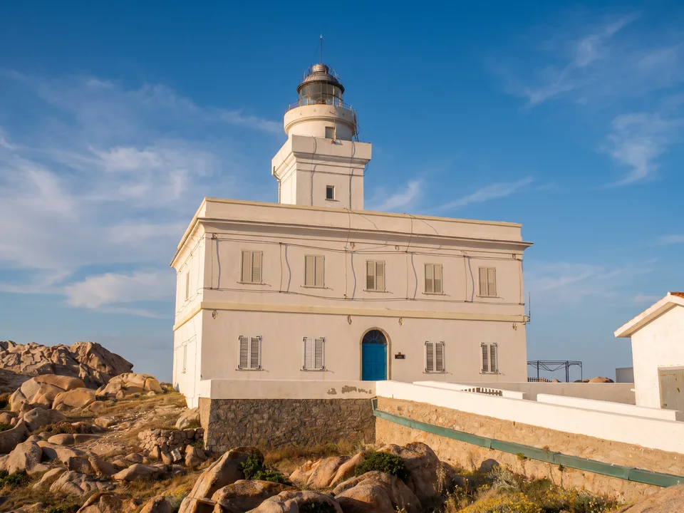 Weißer Leuchtturm von Capo Testa mit blauer Tür zwischen Granitfelsen unter blauem Himmel