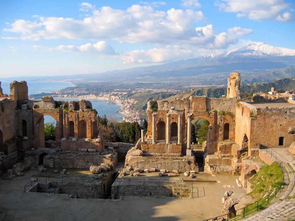 Ruinen des antiken Theaters von Taormina mit Säulen und Bögen, dahinter die Bucht von Giardini-Naxos und der schneebedeckte Ätna