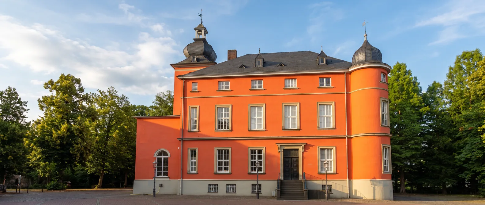 Das rot-orange verputzte Herrenhaus der Burg Wissem mit Zwiebelhaube, rundem Eckturm und weißen Sprossenfenstern im goldenen Abendlicht