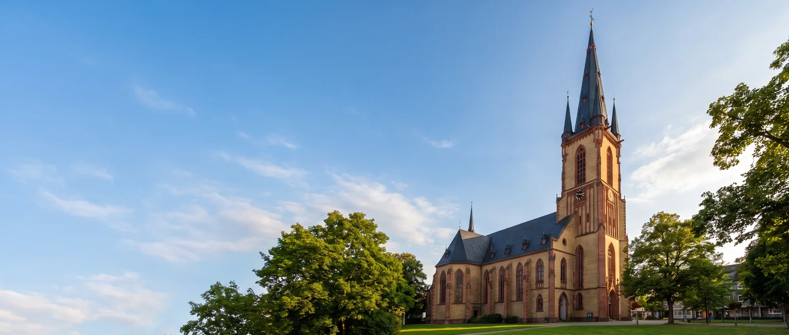 Die neugotische Apostelkirche in Viernheim aus rotem und gelbem Sandstein mit hohem Spitzhelm zwischen grünen Bäumen unter blauem Himmel