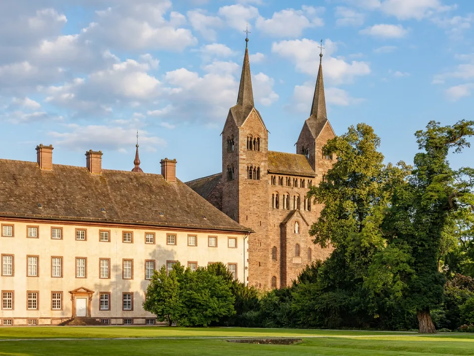 Karolingisches Westwerk mit zwei spitzen Türmen neben dem barocken Schlossflügel von Corvey hinter großen Bäumen im Sonnenlicht