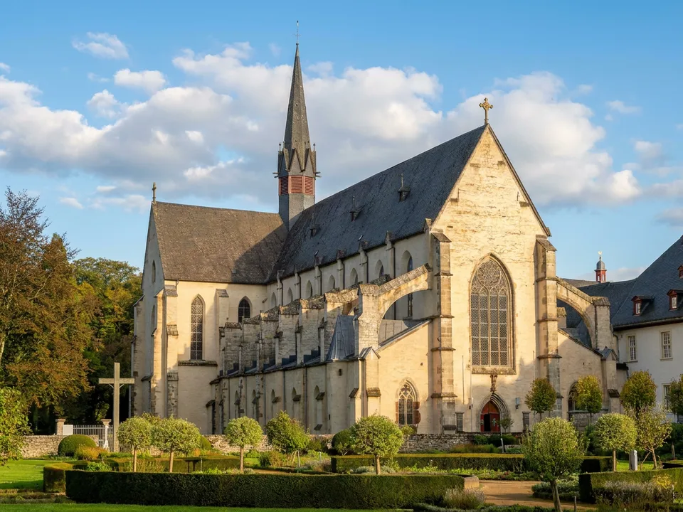 Frühgotische Abteikirche Marienstatt mit hohem Maßwerkfenster, Dachreiter und Strebebögen hinter dem Klostergarten im Sonnenlicht